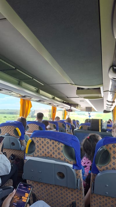 An interior shot captures the passenger seating area of a bus, likely in or near Krivodol, Bulgaria, based on the provided context. The camera is positioned looking forward, showcasing rows of seats with blue and patterned upholstery. The overhead compartment is visible, along with the ceiling and part of the bus windows. Outside the windows, a rural landscape with green fields and a bright sky can be seen, partially obscured by yellow curtains.

Several passengers are seated in the bus. In the foreground, a person's hand holds a smartphone displaying a messaging application with what appears to be a dark, starry background. In the middle ground, various individuals are seated, some with headphones, and their backs are turned towards the camera. Further ahead, more passengers occupy seats, and a child with dark hair and a pink scrunchie is visible.

The overall scene is brightly lit by natural light coming from the windows. The bus appears to be in motion, suggested by the perspective and the outward view. There are no specific landmarks or urban elements to confirm the precise city location beyond the given context of Krivodol, Bulgaria. The atmosphere is casual, indicative of a journey.
