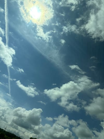 The image shows a bright sun with a halo effect in the upper left quadrant, set against a vast blue sky filled with scattered white and grey clouds. A prominent contrail from an aircraft cuts vertically through the left side of the sky. The lower portion of the image reveals a sliver of lush green trees and vegetation, with visible power lines and utility poles in the far lower left corner. The scene depicts an outdoor environment under a partly cloudy sky, possibly a rural or suburban area, given the presence of trees and utility infrastructure. The specific location in Таймурзино, Russia, is not visually discernible from the provided image alone.
