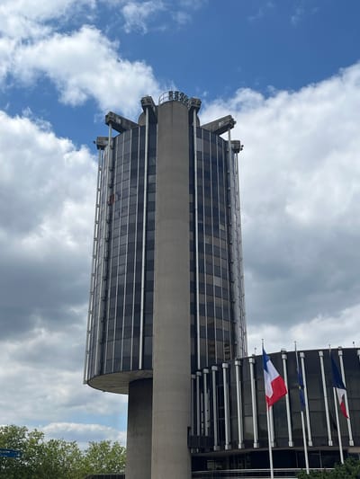 A modern, cylindrical building with a concrete core and a glass facade is visible against a cloudy blue sky. The building has a distinctive flared base and a crown-like structure at its apex. Several French flags and what appears to be an EU flag are flying from flagpoles in front of a lower section of the building, which also features a dark glass facade supported by vertical silver columns. Green trees are visible at the bottom left of the frame. This is likely the Hôtel de Ville (City Hall) of Créteil, France, given its architectural style and the presence of French flags.