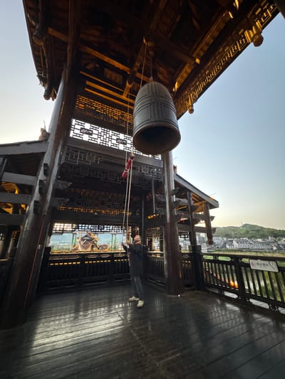 The image shows a large, possibly bronze, bell hanging from a wooden structure in Zhuoshui, China. The structure is part of a multi-story building with intricate woodwork and traditional Chinese architectural elements. A woman stands near the bell, seemingly interacting with the bell's rope. The background reveals the cityscape of Zhuoshui, including a river and hillside. The scene is bathed in the light of early morning or late afternoon, creating a warm and contemplative atmosphere. The floor of the structure is wooden and reflective.