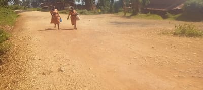 This image depicts a rural or semi-urban scene in Getenga ward, Kenya. Two adult females are walking away from the viewer along an unpaved, reddish-brown dirt road. The woman on the left is wearing a light-colored, possibly orange patterned dress and carrying a blue item in her right hand. The woman on the right, similarly dressed in a light-colored garment, carries a white bag or container in her left hand. The road surface is uneven and contains visible loose gravel and small stones, particularly along the left edge. Green vegetation, including grass and shrubs, lines the left side of the road and appears in patches on the right. In the background, several structures with dark roofs are partially visible, set amidst trees and rolling hills under a bright sky.