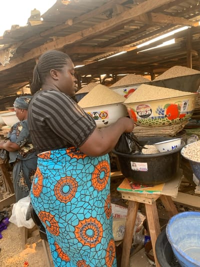 A woman in a gray striped shirt and a vibrant blue and orange patterned skirt stands beside buckets filled with grains under a wooden roof. Her left arm holds a black bag within one of the buckets. Another woman in a patterned headscarf and dress stands slightly behind her. The scene is cluttered, with various containers, buckets, and goods piled up around them.