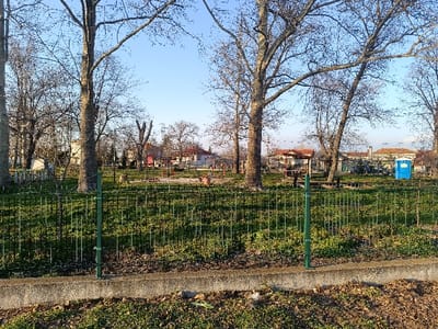 The image displays an unoccupied park or playground area within the city of Izbeglii, Bulgaria, under a clear, light blue sky. In the foreground, a low concrete curb is visible, immediately followed by a green metal fence approximately 1 meter in height, featuring vertical posts and horizontal wire mesh. The ground outside the fence consists of sparse green grass and scattered dry leaves, while the area within the fence is predominantly covered in green grass with some visible soil patches.

The midground is characterized by several tall, deciduous trees with bare branches and thick trunks, suggesting late autumn or winter. A children's playground is situated among these trees, featuring structures such as red-seated swings, a red/orange slide, and other climbing equipment. To the right of the playground, a wooden gazebo with a reddish-brown roof is present, and further to the right, a bright blue portable toilet is visible.

In the background, multiple low-rise buildings with light-colored facades and terracotta-tiled roofs are observed beyond the park boundaries. No individuals are present within the frame.