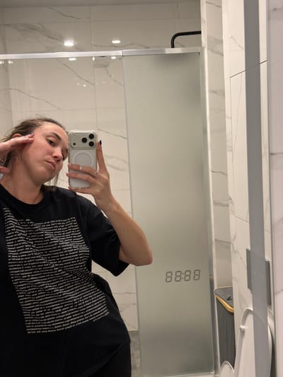 A woman in a black t-shirt takes a selfie in a modern bathroom. She is wearing rings on her fingers and has blue nail polish. The bathroom features marble tile walls, a glass shower enclosure, and a digital clock on the shower door displaying "88:88". A trash can is visible behind the shower door. The scene is indoors and appears to be a domestic setting. The location is not specified in the image or metadata.