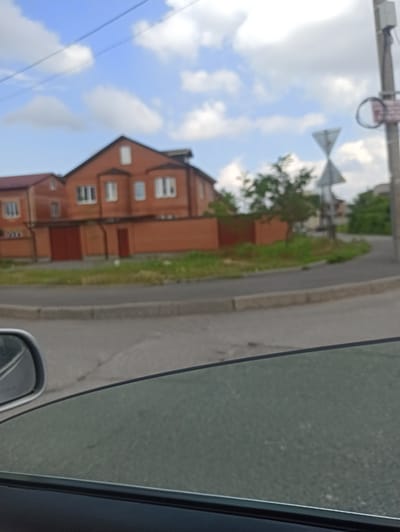 The image depicts a street scene in Verkhnii Lars, Russia, with a focus on a brick house and surrounding elements. The house is a multi-story structure with a red brick facade and a dark-colored roof. It features multiple windows and a large garage door. A red brick wall surrounds the property. In the background, there are trees and other buildings, indicating a residential area. A street with a curb is visible in the foreground, with a car's side mirror and tinted window suggesting the photo was taken from inside a vehicle. Traffic signs, including a triangular sign, are present on a utility pole on the right side of the image. The sky is partly cloudy with a blue hue. The scene appears to be captured during daylight hours.