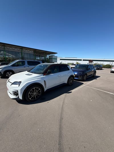 The image captures an outdoor parking lot under a bright, clear blue sky, likely in Aubange, Belgium. Several cars are parked on a gray asphalt surface marked with white lines.
In the foreground, a modern white SUV or crossover with black accents is prominently featured. It has distinctive black and silver multi-spoke alloy wheels, black wheel arch trim, and sharp, contemporary front lighting. A subtle red trim is visible along its roofline. Behind this vehicle, a dark blue car, possibly another SUV or a large hatchback, is parked, with its front lights illuminated. Further back, other cars are partially visible.
To the left of the parking area, a modern building with large glass panels and a visible metal framework extends across the scene. Some greenery, including bushes and small trees, lines the base of the building. The strong sunlight indicates it is daytime, likely midday or early afternoon, casting distinct shadows from the cars onto the ground. The sky is entirely cloudless.
On a white sign located on the building in the background, faint text that appears to read "jardinerie" is visible, suggesting the building may be a garden center or part of a commercial complex. No people are present, and the primary activity depicted is the parking of vehicles.