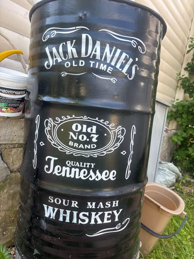 An outdoor scene in Лотос Снт (марьино), Russia, features a large, black barrel painted with the Jack Daniel's Old No. 7 Tennessee Sour Mash Whiskey logo. The barrel is positioned vertically and appears to be outdoors, possibly in a yard or garden, with a light-colored wooden wall visible in the background. A tan plastic bucket is placed next to the barrel on the right. In the foreground and to the left, there are some green plants and a stone wall. A white plastic container with a yellow lid and some writing is also visible to the left of the barrel. The overall impression is of a rustic, outdoor setting.