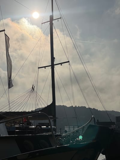 The image captures a silhouetted sailboat mast and rigging against a hazy sky with the sun partially obscured by clouds. The sun creates a bright halo effect. A flag is visible hanging from the left side of the mast. The foreground shows the upper deck of another boat with metallic railings and what appears to be a steering wheel. The name "MAZTUR" is visible on the side of this boat. In the background, a tree-covered hill or mountain range forms a silhouette against the sky, suggesting a coastal or waterfront location. The overall atmosphere is slightly overcast and diffused light. Given the context of Beşiktaş, Turkey, this scene likely depicts a harbor or marina with traditional Turkish Gulet-style boats, common in the region.