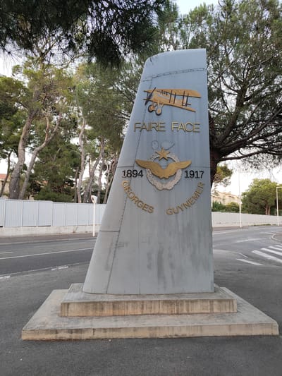 This media file shows a monument in Perpignan, France, dedicated to Georges Guynemer, a famous French aviator. The monument is shaped like the tail fin of an airplane, made of grey metal with rivets. On its surface, gold lettering spells out "FAIRE FACE", "1894", "1917", and "GEORGES GUYNEMER". Above the name, there's a depiction of a biplane and an insignia featuring wings and a star. The monument stands on a concrete base on the side of a road, with a pedestrian crossing visible in the background. Trees, some with pine needles, surround the monument, and a white fence runs along the roadside. The scene appears to be set during the daytime, under overcast or clear skies. The overall impression is one of remembrance and tribute to a historical figure.