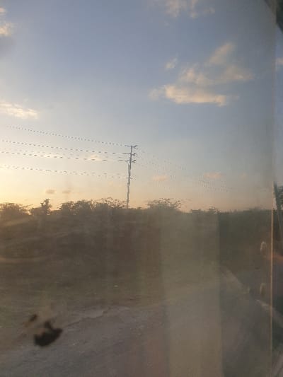 A view of a light blue sky with fluffy white clouds and a power line is visible. A large utility pole stands in the frame, supporting multiple power lines stretching across the sky. Vegetation lines the horizon. The image's right portion presents blurred, reflective surfaces, hinting at the image being captured through a window or similar barrier. A dark smudge is visible in the lower right corner of this reflective surface.