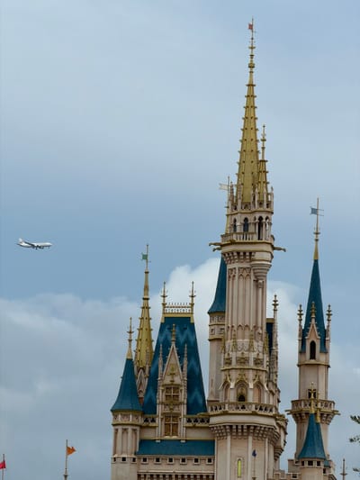 An airplane is visible in the sky to the left of a fairytale-style castle. The airplane is white with red and blue markings. The castle has multiple spires, some of which are gold and some are blue. The castle is an iconic landmark of Tokyo Disneyland, located in Urayasu, Chiba Prefecture, Japan. The sky is overcast and cloudy.