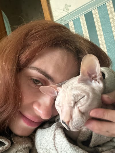 This is a close-up, portrait-style photograph of a woman and a Sphynx cat. The woman has auburn hair and is looking at the camera with a slight smile. Her left eye is visible, with brown irises and defined eyebrows. Her lips are painted with a pink lipstick. The Sphynx cat, with its characteristic hairless skin and large ears, is nestled against her face, its eyes closed as if sleeping or content. A hand is gently petting the cat's head.

The background consists of striped wallpaper with alternating bands of light blue and cream, featuring a subtle, repeating pattern. To the left, a portion of a wooden surface is visible, possibly furniture. The overall lighting suggests an indoor setting, likely during the daytime. The mood of the image is intimate and affectionate, highlighting the bond between the woman and her pet. There is no visible text in the image. The location context is Togliatti, Russia.