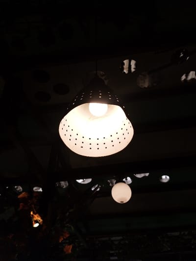 A dimly lit indoor scene features two visible light fixtures suspended from the ceiling. The primary fixture is a perforated, conical lamp emitting a bright, diffused glow. Below it, a smaller, spherical lamp provides a softer, less intense illumination. Dark structural elements and ambiguous foliage obscure the background, creating a contrast with the glowing light sources. The overall atmosphere is intimate and subdued.