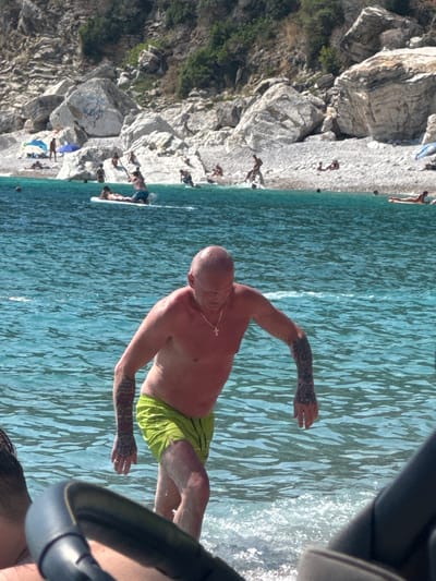A man with a shaved head and tattoos on his arms is standing in the clear, blue water of a beach. He is wearing bright green swim trunks and a thin necklace. The sun is shining, casting bright highlights on the water's surface, suggesting it is daytime and the weather is warm and sunny.
Behind the man, a rocky, pebble beach slopes down to the water. Several other people are visible on the beach, some lying down and others standing. A few dark spots in the water might be people swimming or rocks. The scene is set against a backdrop of large, grey rocks and some green vegetation on a hillside. The water is calm with gentle ripples and some small waves breaking near the shore. The perspective of the image suggests it was taken from a slightly elevated position, possibly from a boat or a higher point on the shore. The overall impression is a peaceful day at a scenic, Mediterranean-style beach.