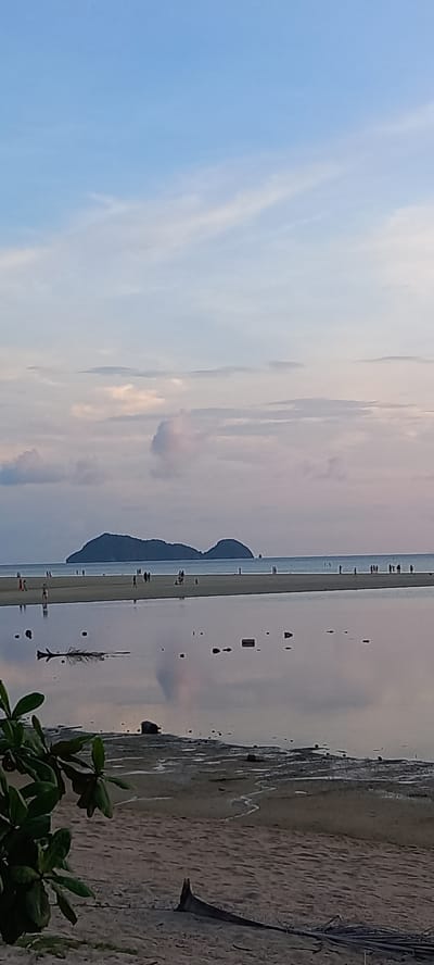 The image shows a coastal scene at Baan Sri Thanu, Thailand, featuring a long, narrow sandbar extending into the sea. The sky is partially cloudy. Several people are seen walking along the sandbar. Two islands are located on the sandbar that are close to one another. Reflections of these features are visible in the water. The right side of the image includes a sandy beach area with some vegetation in the foreground. Debris is also visible on the beach.