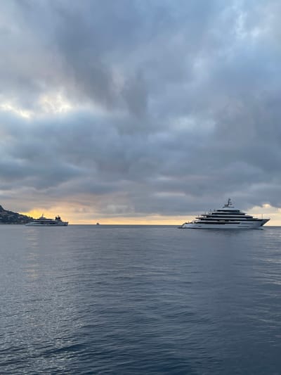 The image depicts a serene seascape with a dramatic sky. Two large yachts are anchored in the calm, dark blue water. The yacht in the foreground is a multi-decked vessel with a white hull and numerous windows. The second yacht is smaller and farther in the distance, positioned towards the left side of the frame. A coastline with buildings and lush vegetation is visible on the far left, suggesting a proximity to land. The sky is overcast with heavy, gray clouds that are broken in the middle by a band of soft, golden light, indicative of either sunrise or sunset. The overall mood of the image is tranquil and somewhat somber due to the cloudy sky, but the sliver of light offers a touch of hope. The location is the coastal area near Èze, France, known for its picturesque views of the Mediterranean Sea.