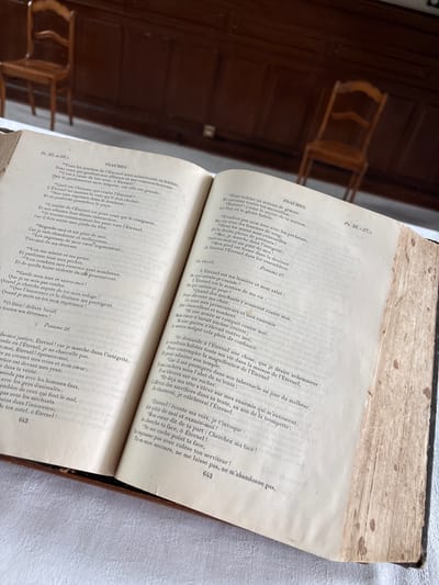 The image shows an open, aged book on a white textured surface, likely a tablecloth. The book is bound in dark material, and its pages are yellowed and stained with age. The text appears to be in French, with "PSALMES" prominently displayed. The left page contains Psalm 25, with verses numbered. The right page features Psalm 26 and Psalm 27, also with numbered verses. The book is positioned at an angle, revealing part of its spine and the texture of the paper. The background is blurred but suggests an indoor setting with wooden furniture, possibly chairs, hinting at a historical or ecclesiastical environment. There are no people or any discernible action taking place in the image.