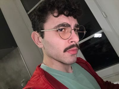 A person with curly dark hair, wearing round glasses and sporting a mustache, is angled slightly towards the right. They are wearing a light green t-shirt and a red jacket. A window is visible behind them, reflecting a light source. The background is mostly white, and the lighting is slightly dim.