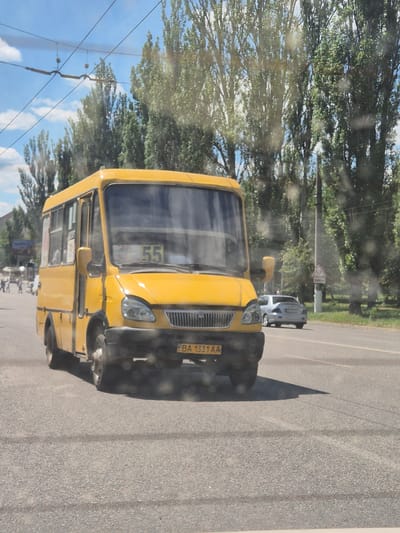 The image shows a yellow bus on a paved road, likely in Kropyvnytskyi, Ukraine, given the context. The bus is the primary object in the foreground, viewed from the front passenger side. It is a large vehicle with prominent headlights and a license plate that reads "BA 1331 AA". The number "55" is displayed on the front of the bus, possibly indicating its route or designation.
In the background, there are trees lining the road, suggesting an outdoor setting, possibly a suburban or rural area. A light-colored car is visible further down the road, behind the bus, and a pedestrian crossing sign is also present. The sky is blue with some clouds, indicating daytime. The overall lighting suggests it's a sunny day. Reflections on the bus's windows, particularly the windshield, hint that the photo might have been taken from inside another vehicle, perhaps a passenger in a car or another bus. There are no visible people in the immediate vicinity of the bus, though the presence of a car and traffic signs implies a public road with ongoing activity. The scene appears to be captured during daylight hours.