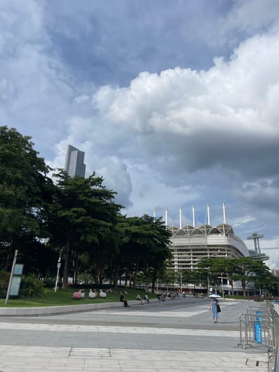 The image captures a view of the Yuexiu District in Guangzhou, China. The scene features a modern stadium with a distinctive white, tent-like roof structure supported by prominent poles. To the left of the stadium, a tall, angular skyscraper rises into a sky filled with dramatic, dark clouds. In the foreground, a tree-lined park area with green grass and scattered ornamental sheep sculptures is visible. Several people are seated on a low wall in the park, and others are walking or standing along a paved walkway. A woman in a blue dress walks across the walkway carrying a white umbrella. In the distance, a tall tower with a platform is visible. The overall atmosphere is overcast, suggesting a possibly stormy day.