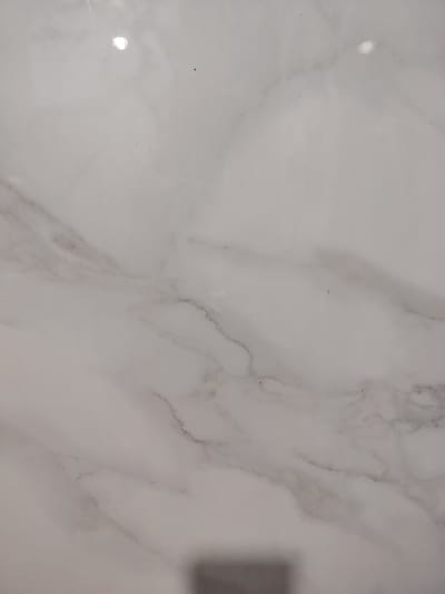 The image displays a close-up view of a white marble surface with subtle grey veining. The texture appears smooth and polished, with a reflective quality that creates bright highlights. There are a few small dark specks scattered across the surface, possibly dust or imperfections. The lighting suggests a well-lit indoor environment, likely artificial. No specific location in Pitnak, Uzbekistan can be identified from this image, as it focuses solely on the material's pattern and texture. The image does not contain any people, actions, or interactions.