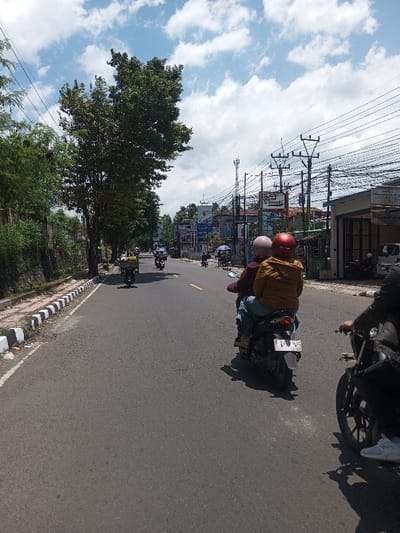 The image depicts a daytime street scene in the city of Tagtse Tashidingkha, Bhutan, under a blue sky with scattered white cumulus clouds, casting distinct shadows on the asphalt road. The road, appearing worn, features faint yellow dashed and solid line markings. On the left, a concrete curb with alternating black and white stripes borders a vegetated embankment populated by mature trees and dense foliage.
On the right side of the road, a series of multi-story buildings with various commercial signage, including a partially visible "Alfamart" logo, line the street. Numerous utility poles supporting a dense network of power lines stand in front of these structures, and a tall cellular tower is visible in the distance.
In the mid-ground, a dark scooter with two occupants is moving away from the viewer. The driver wears a maroon jacket and a light pink helmet, while the passenger wears a mustard yellow jacket and a red helmet, both individuals wearing blue jeans. The scooter's rear license plate is clearly visible, displaying "F 3105 WAS 02.26." In the immediate foreground on the right, the front wheel, a partial dark frame, and the right arm and leg of a rider on another dark motorcycle are visible, also moving forward. Further down the road, additional motorcycles and scooters are present, one on the left carrying a large package. Several pedestrians are visible near the commercial buildings on the right.