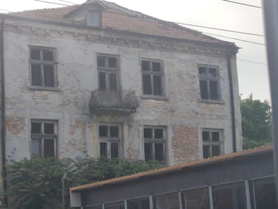 This image depicts a two-story residential building in a state of advanced disrepair, located in Lom, Bulgaria. The facade is characterized by severely deteriorated light-colored plaster, which has extensively peeled and crumbled, exposing sections of red brick masonry beneath. The building features multiple rectangular window openings on both visible floors, fitted with multi-pane wooden frames. The window panes appear dark or absent, suggesting either broken glass or shadowed interiors. Centrally located on the second floor is an ornate, curved wrought-iron balcony. The roof is composed of red ceramic tiles and includes a gabled dormer window with a single pane near the peak. A deteriorated cornice and decorative band are visible below the roofline. Dense green foliage partially obscures the lower facade of the main building and the roofline of a lower, separate structure in the immediate foreground. This foreground structure has a low-pitched roof with a light-colored fascia and appears to have large windows, indicating a different architectural style or function. Overhead utility lines are visible traversing the upper portion of the frame against a pale, overcast sky. A dark vertical column, possibly a utility pole or building edge, frames the right side of the image. No individuals or discernible activity are present within the scene.