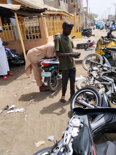 The image depicts an outdoor motorcycle repair or sales scene in Fagge D2, Nigeria, under bright daylight. In the foreground, a black motorcycle lies on its side on unpaved, dusty ground scattered with debris. To the left, a man in a light brown traditional garment and dark sandals leans over the seat of an upright grey and black motorcycle featuring a visible Nigerian license plate. Next to him, a young male in an olive-green t-shirt, dark trousers, and open-toed sandals, with dark sunglasses resting on his head, stands facing right and examines a small, dark object held in both hands. To the right of these individuals, several disassembled motorcycle frames and parts, including wheels and engine components, are scattered on the ground. In the mid-ground, a single-story building with a yellow metal fence and a corrugated awning is visible. The background reveals a paved street with multiple motorcycles, a blue car, pedestrians, and streetlights, indicating a bustling urban environment with multi-story buildings lining the road.