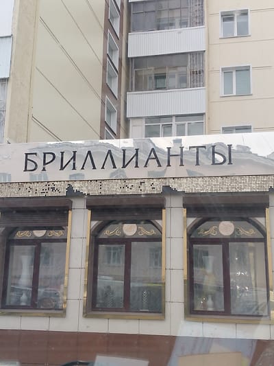 This image shows the exterior of a building with a sign that reads "БРИЛЛИАНТЫ" (Brilliants) in large, dark, serif font. The sign is mounted on a reflective surface, creating a mirror-like effect. Below the sign, there is a decorative band made of small, circular metallic elements, resembling sequins or LEDs.
The building has a light-colored facade with dark-framed windows. Three windows are prominently visible in the foreground, each with arched tops and ornate gold trim. Inside the windows, reflections show some interior elements, possibly vases or decorative items, along with further reflections of the building's exterior. The windows appear to be covered by sheer curtains.
The architecture suggests a commercial establishment, likely a jewelry store given the sign's text. The overall impression is one of a moderately formal or elegant storefront. The lighting suggests it might be daytime, but the reflections and the quality of the light make it difficult to determine the exact time of day or weather conditions. The location context provided, Magadan, Russia, implies a northern climate, which could influence architectural styles and the general appearance of the scene. No people are visible in the image.