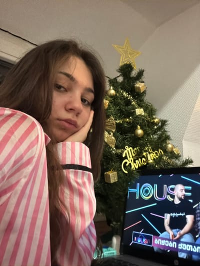 A woman in pink striped pajamas rests her head on her hand. A Christmas tree adorned with gold ornaments stands behind her. A gold star tops the tree, and a gold inscription wraps around its center. To the right, a television screen displays a show called "HOUSE." A man's face is visible on the screen, along with Georgian text. The scene suggests a cozy, relaxed atmosphere indoors.