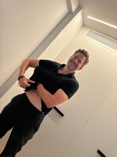In what appears to be a modern, minimalist interior, likely a domestic or hotel setting, a Caucasian man with blond hair and a light complexion is pictured. He is wearing a black polo shirt and black pants. His shirt is pulled up, revealing a portion of his abdomen. He is looking at the camera with a smile. His left arm is bent and appears to be holding the shirt up, while his right arm is bent at the elbow. On his left wrist, he wears a black watch, and on the same wrist, a dark woven bracelet. The environment features plain white walls and what seem to be built-in cabinets or doors, one with horizontal ventilation slits. A linear light fixture is visible on the ceiling, suggesting contemporary architectural design. The lighting is bright and casts soft shadows. There is no visual information in the image to suggest this scene is located in Murcia, Spain.
