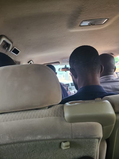 The image shows the interior of a vehicle, likely a van or SUV, from a rear-facing perspective. Three individuals are visible, all appearing to be adult males. The driver, or front passenger, is obscured by the seat in the foreground. The primary focus is on the heads and upper bodies of two men seated in the middle and rear rows. The man in the middle row, closer to the camera, has short black hair and is wearing a dark blue jacket or shirt. The man in the rear row, to his right, has a checkered shirt. The interior of the vehicle has a light-colored headliner and fabric-covered seats. A rearview mirror is visible, reflecting the exterior, which suggests daylight and possibly an urban or suburban environment. There is a light fixture on the ceiling of the vehicle. The overall impression is of individuals traveling within a city.