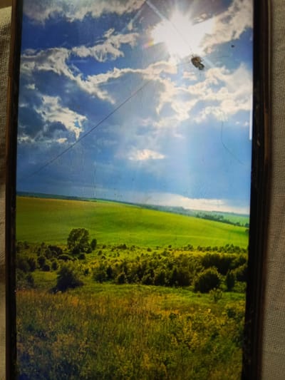 The image displays a scenic view of rolling green hills under a dramatic sky. Sunlight streams through a break in the clouds, illuminating the landscape. There are trees and dense foliage in the foreground and middle ground. A thin, dark line, possibly a wire or a scratch on the screen, stretches across the upper portion of the image. The overall scene is a natural landscape with no man-made structures or identifiable landmarks suggesting a specific location. No people or actions are depicted in the image.