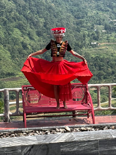 A person dressed in vibrant traditional attire is captured posing outdoors against a mountainous backdrop in Thông Nguyên Commune, Vietnam. The subject, likely a woman, stands on a stone-tiled platform, holding out the voluminous, bright red pleated skirt of her dress.

She wears a richly patterned top, possibly a vest or short-sleeved garment, adorned with intricate designs. Her head is crowned with an elaborate, tall traditional headpiece featuring red fabric and prominent white decorative elements. Visible tattoos adorn her arms, and she is barefoot.

Behind the subject is an ornate red metal structure, resembling a bench or railing, with intricate cut-out patterns that suggest traditional Vietnamese motifs or calligraphy. On this structure, Vietnamese text is visible: "NỘI LƯU GỬI NẾN SĐT: 0948".

The background is dominated by lush, dense green mountains and valleys, characteristic of Vietnam's highland regions, with possible hints of terraced fields on the slopes. The foreground features a pathway or platform constructed from dark grey and white striped stone tiles, bordered by small natural rocks.

The image is well-lit by clear daylight, suggesting a sunny day, likely midday or early afternoon, with sharp shadows. The overall scene emphasizes cultural heritage set against a majestic natural landscape.