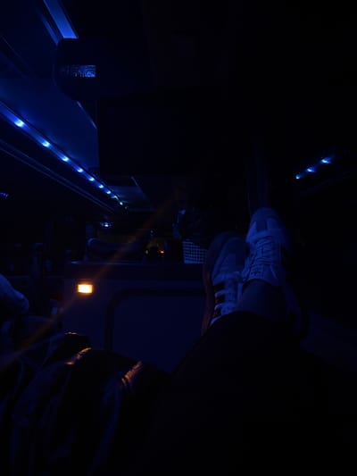 A person sits, legs crossed, in a dimly lit interior with blue lighting. The individual wears white Adidas sneakers. A metal safety barrier runs along the side. An orange light is visible in the background. The image suggests a confined space, possibly a train or vehicle interior at night.
