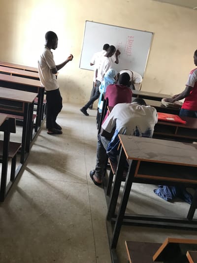 The image, originally rotated ninety degrees clockwise, portrays an indoor educational setting, likely a classroom or study area in Ilorin, Nigeria. Several young men, presumably students, are engaged in a learning activity centered around a whiteboard.

In the foreground, a student in a white shirt and black trousers stands facing the whiteboard, with his right arm slightly raised, appearing to observe or participate in a discussion. Immediately behind him, a group of three students is actively writing or drawing on a large white whiteboard. One student wears a light blue top, while the others are in white shirts.

On the right, other students are situated at dark-framed desks with light-colored wooden tops. One student wears a red and white striped top, while another has a white t-shirt with faint blue text on the back, possibly a university logo. A red bag is visible among the desks. The floor is light grey, appearing to be speckled concrete or tile. The walls are a plain, light beige color.

The whiteboard displays text written in both red and black markers. Clearly visible red text includes a numbered list: "1. Diagram", "2. Infernal", "3. Music", "4. Prayer", "5. Traffic", "6. Anima", "7. Science", "8. Speech", and "9. Factor". Other black text on the board is less distinct but appears to be additional notes or headings.

The scene suggests a focused academic environment, with students collaborating or presenting information. The attire, including sandals, is consistent with the Nigerian context. The lighting is artificial, characteristic of an indoor setting, and does not indicate the time of day.