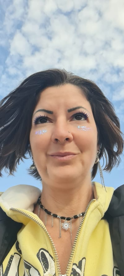 A woman is looking down at the camera from a low angle in an outdoor setting with a partially cloudy blue sky as the background. She has brown hair, light skin, and is wearing makeup that includes glittery accents near her eyes and dark mascara. A silver nose ring is visible in her left nostril.

She is wearing a yellow hoodie with a partially zipped-down front, revealing a silver necklace with black beads and a pendant shaped like a star. Silver chains also dangle from her earrings. The picture gives the impression that she is somewhere where the weather is nice and the sky is visible. No specific details confirm that the location is in Nahariyya, Israel.