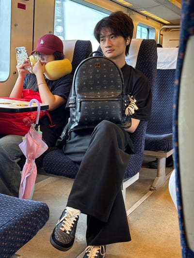 On a train, an individual with dark hair and a black t-shirt sits next to a window and wears black pants. An MCM backpack rests on their lap. Beside them, another person, wearing a maroon baseball cap and a yellow neck pillow, looks at a mobile phone. A partly open multi-colored umbrella rests beside them. The train seats are upholstered in a blue patterned fabric. The location is the city of Baiyun District, China.