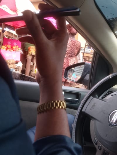 The image depicts an interior view from a vehicle in Aketu-Oja, Nigeria, likely on a street market day given the presence of vendors and goods visible through the car window. In the foreground, a person's arm, adorned with a gold watch, is positioned to hold what appears to be a smartphone. This arm is resting on the car's interior paneling. Outside the vehicle, a woman in a leopard-print dress and headwrap is seen standing near a market stall. Behind her, a variety of goods, including pineapples and what might be oranges or peaches, are displayed on tables and in crates. The market appears to be set up under a pink awning. A side mirror of the car reflects a child wearing a blue and pink shirt, and a portion of the market stall. The steering wheel of the vehicle is partially visible in the lower right portion of the frame, showing the Nissan logo and airbag indicator. The scene suggests a busy, outdoor market environment typical of a Nigerian town.