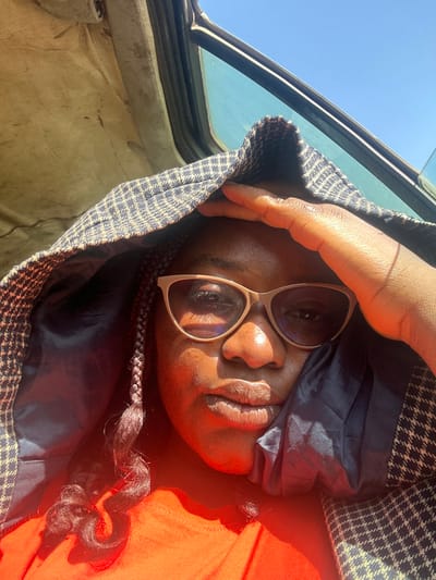 An adult female is depicted in a close-up, seated inside a vehicle under direct sunlight in Rimau, Nigeria. She has dark skin, wears light-colored framed eyeglasses, and an orange top. Her hair is braided, with some dark, curled braids visible on her left side. She holds her right hand to her forehead, partially shielding her face. A dark-colored fabric with a subtle checkered pattern is draped over her head like a hood, its dark blue interior lining visible. Above her, the textured, light-colored headliner of the vehicle is visible. A vehicle window to her right shows a clear blue sky. Strong sunlight illuminates the scene, casting highlights on her face, particularly her right cheek and chin, and on her glasses. She looks directly forward.