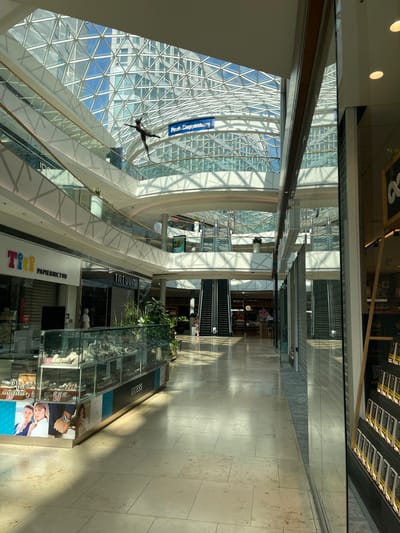 The image shows the interior of a modern shopping mall with a large, glass-paneled, geometric-patterned ceiling that allows natural light to flood in. Suspended from the ceiling by thin wires is a metallic sculpture of an acrobatic figure in mid-air. The mall features multiple levels with white-paneled balconies and escalators visible in the background. On the ground floor, shops line the wide, tiled walkway. To the left, a jewelry store display case showcases watches and other accessories. A "Tiff" stationery store and a "Tatuum" clothing store are also visible. Further down the walkway, escalators lead to the upper levels. A person is seen walking on the right side of the walkway. The mall appears to be the Eurovea shopping center in Bratislava, Slovakia, given the signage for "Pestloppensburg," which is an older name for the area and likely a reference in the mall's context.