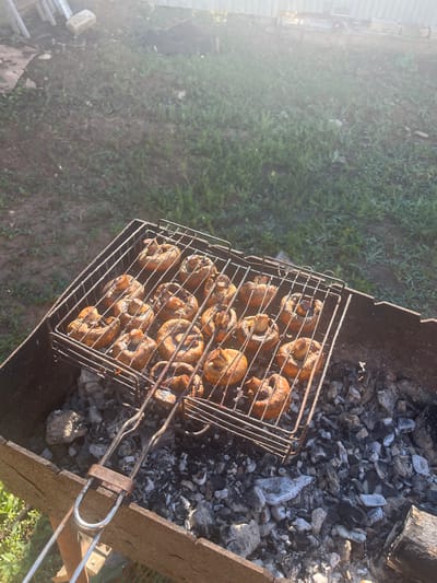 The image shows whole mushrooms being grilled on a barbecue. The mushrooms are placed on a metal grill rack, which is positioned over hot coals in a barbecue pit. The scene is set outdoors, likely in a backyard or park, with green grass and a fence visible in the background. The sunlight suggests it is daytime, possibly late afternoon or early evening due to the warm, soft light and shadows. The mushrooms appear to be cooked, with some charring on their surfaces, indicating they are being prepared for consumption. The overall mood is one of casual outdoor cooking and enjoyment. No people are visible in the image. There is no visible text. The location context is Rakhinka, Russia.