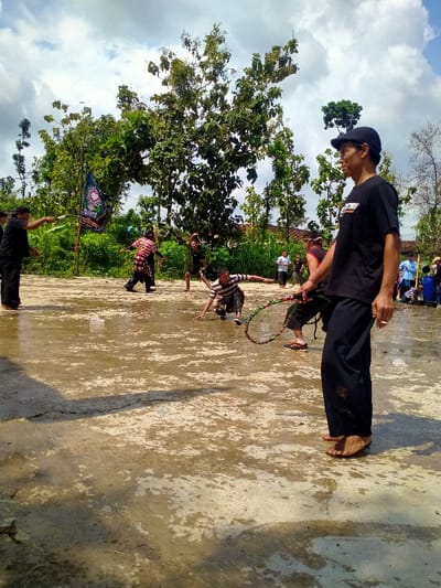 The image shows a group of men in a clearing in a wooded area in Kecamatan Kedawung, Indonesia (latitude: -7.518, longitude: 111.029). The men are gathered in a muddy clearing. Some of the men are practicing a martial art, while others are standing around watching. One man in a black t-shirt and black pants is holding a long rope and standing near the edge of the clearing. He is watching the men practice. There is a large tree with leafy branches in the background. There is also a flag hanging from a pole in the background.  The ground is covered with mud and the sky is mostly clear.