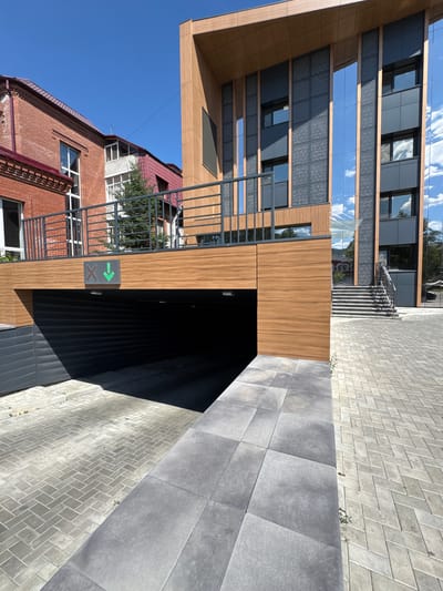The image shows the entrance to an underground parking garage in Blagoveshchensk, Russia. The garage entrance is a dark opening with a ramp leading down to it. Above the entrance, there is a wooden facade with a sign displaying a red "X" and a green downward arrow, indicating entry is permitted. To the left of the garage is a brick building with red roof tiles and several windows. To the right is a modern building with a glass facade and wooden accents, featuring a staircase leading up to the building's entrance. The ground is paved with interlocking grey bricks. The sky is clear and blue with some scattered clouds. No people are visible in the image.