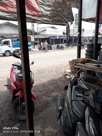 The image shows a street in Kecamatan Haurgeulis, Indonesia (latitude: -6.415, longitude: 107.958) viewed from a narrow alley. Two motorbikes are parked in the foreground. A red motorbike is on the left and a black motorbike is on the right. The alley is lined with buildings, including a white building with a sign that says "SAMSAT". There are several people walking on the street, including a man wearing a white shirt and blue pants who is walking towards the right side of the image. The street is paved with a gray, gravel-like material. The image is taken from a slightly elevated position, as the viewer is looking down onto the street.