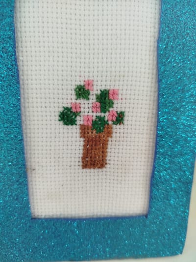 The image shows a close-up of a cross-stitch embroidery featuring a small potted plant with pink flowers and green leaves. The embroidery is on white Aida fabric and is framed by a textured, glittery blue material. The background is out of focus. The specific location in Крывасельскі сельскі Савет, Belarus is not discernible from the image.
