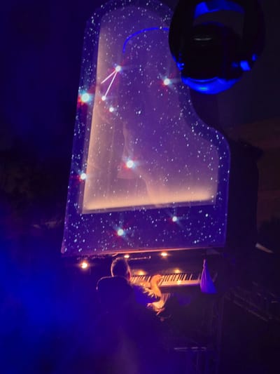 A grand piano, decorated with a starry night sky projection, sits on a stage. A person is playing the piano, their hands visible on the keys. The stage is bathed in blue and purple light, with some spotlights illuminating the scene. The piano's lid is open, revealing the keys and a portion of the interior, which is also lit. The overall impression is of a theatrical performance or concert, possibly with a celestial theme. The image is dark, with the lighting contributing to a dramatic and ethereal atmosphere. The crops provide close-ups of the piano's projected surface, the person's hands on the keys, and the surrounding stage lighting.