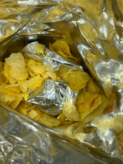 A close-up, overhead shot shows a bag of potato chips, likely in a shopping context. The bag is made of reflective foil, with some crumpled sections reflecting light and casting shadows. The chips are golden yellow and appear to be broken into various sizes. The scene is set in Stepantsminda, Georgia, a location renowned for its natural beauty and mountainous terrain, but this image does not show any of those features.