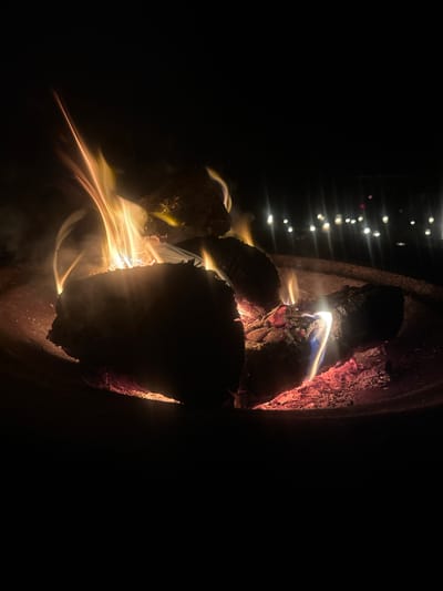 The image depicts a close-up view of a bonfire at night. The central subjects are several pieces of burning wood, with flames of yellow and orange licking upwards. Embers and ash are visible in the fire pit, glowing red and orange. Wisps of smoke rise from the fire. In the background, out of focus, are numerous small, bright lights, suggesting a distant gathering or settlement, possibly in the outer areas of Darkwood, Australia. The overall environment is dark, emphasizing the illumination provided by the fire. There are no people or animals visible in the frame.