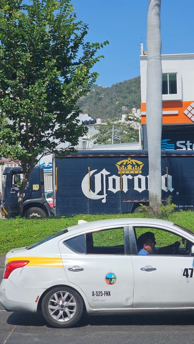 The image shows a white taxi with the number 47 on its roof, displaying the license plate "A-525-FNX" and a government seal, suggesting it is a licensed cab. The driver is visible through the window. Parked next to the taxi is a large, blue Corona beer truck, indicative of commercial activity. In the background, there is a building painted with orange and white stripes, as well as a mountain that suggests the location is near hilly terrain. The lush green foliage and clear sky indicate a sunny day, potentially in a tropical or subtropical climate. Based on these observations, the image is likely taken in Acapulco, Mexico, a city known for its hilly coastal landscape and commercial activity.